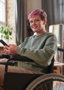 a young woman with short pink hair in a wheelchair at a work desk, holding a mobile phone.